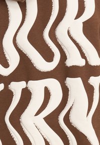 Paceltas baltas burti "R" un "K" ar teksturētu malu uz brūnas auduma, veidojot drosmīgu, viļņotu tipogrāfisku rakstu.