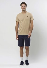 Beiges T-Shirt mit V-Ausschnitt und kleinem gestickten Logo, kombiniert mit marineblauen Shorts. Das Model trägt weiße Socken und dunkle Sneaker und steht vor einem hellen Hintergrund.
