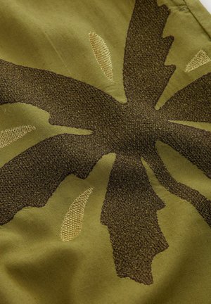 Green fabric with a textured leaf pattern in dark brown and gold thread. The design includes raised embroidery and subtle variations in shade.