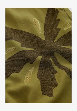 Tissu vert avec un motif de feuilles texturé en marron foncé et fils dorés. Le design inclut des broderies en relief et des variations subtiles de teinte.