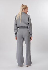 Femme avec queue de cheval blonde portant une veste courte grise à manches longues et un pantalon large assorti, debout et de dos.