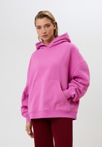 Felpa oversize rosa con tasca frontale, polsini a costine e una texture uniforme. Presenta un cappuccio con cordino e un branding sottile sul petto.