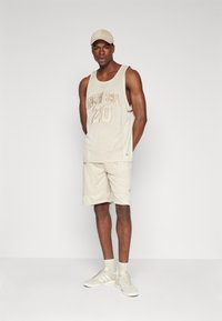 Débardeur en maille beige avec le texte "20" et "NEW ERA", associé à un short assorti. Porté avec une casquette assortie et des chaussures de sport beiges.