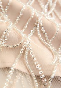Beaded fabric showing intricate patterns with clear and silver beads on a soft nude background, featuring smooth and textured areas.