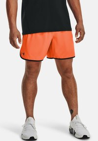 Orange idrottsshorts med svarta detaljer, gjorda av lättviktigt material. Bärs med en svart topp och vita sneakers. Visar synlig ben-tatuering.