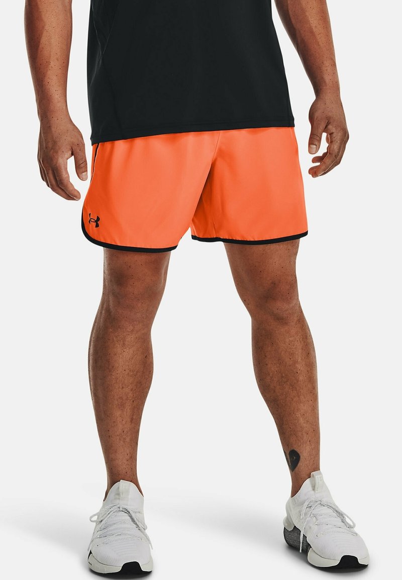 Orange idrottsshorts med svarta detaljer, gjorda av lättviktigt material. Bärs med en svart topp och vita sneakers. Visar synlig ben-tatuering.