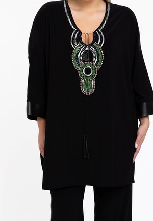 Tunique noire avec encolure géométrique ornée de perles vertes, blanches et beige, liens à pompons et manches trois-quarts avec poignets en cuir.