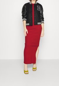 Schwarze Collegejacke mit Lederärmeln und gestreiftem Rippenstrick, getragen über einem figurbetonten roten Kleid mit einem seitlichen Schlitz, kombiniert mit grünen offenen Sandalen.