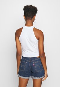 Hvit ermeløs crop topp med høy nakke, sammen med mørke denimshorts med oppbrettede kanter og fremtredende baklommer.