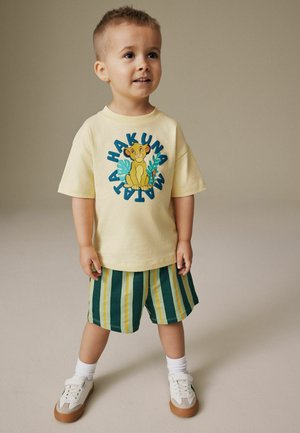 Junger Junge steht da, trägt ein gelbes "Hakuna Matata"-Shirt mit Simba und grün gestreifte Shorts, weiße Socken und Turnschuhe.