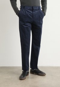 Pantalon bleu marine au design tailleur, avec des plis à l'avant. Porté avec des baskets noires et un pull gris ajusté.