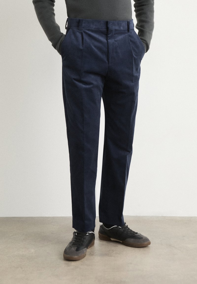 Pantalon bleu marine au design tailleur, avec des plis à l'avant. Porté avec des baskets noires et un pull gris ajusté.