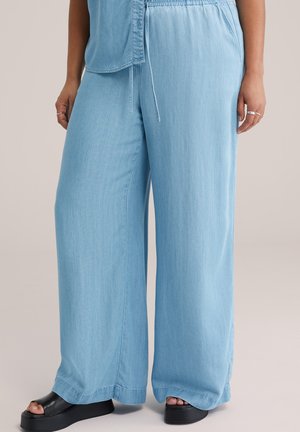 Kvinde iført lyseblå vidde bukser med elastisk talje og sorte platform-sandaler med åbne tæer mod en neutral baggrund.