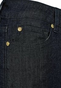 Tamni traperice s prikazom materijala iz bližeg ugla. Istaknuti detalji uključuju mjedenu opremu i šavove duž džepova.