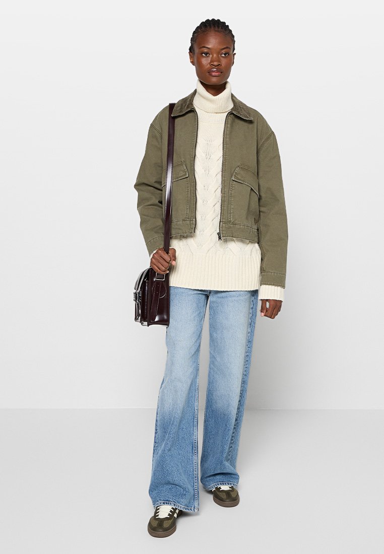 Olivgrüne kurz geschnittene Jacke mit Fronttaschen, cremefarbener Zopfmuster-Pullover, blaue Schlagjeans, braune Sneakers und eine dunkle Umhängetasche.