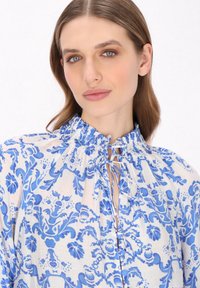 Langärmlige Bluse mit einem weißen Hintergrund und filigranen blauen Blumenmustern, einem gerafften Ausschnitt und dezenten Bindedetails an der Vorderseite.