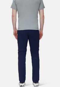 Man som bär en grå t-shirt med kort ärm och marinblå zip-off vandringsbyxor, stående med ryggen vänd framåt, mot en vit bakgrund.