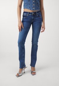 Mujer con jeans ajustados de color azul, top de mezclilla sin mangas y corto, y sandalias de tacón alto plateadas, de pie contra un fondo sencillo.