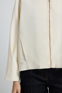 Beige ritsjack met een gestructureerd oppervlak en lange mouwen, gecombineerd met donkerdenim jeans met contrasterende stiksels.