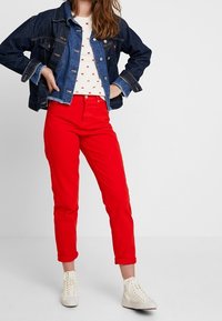 Jeansjacke mit Knopfleiste, rote Baumwollhose und weiße Turnschuhe. Das Oberteil hat ein Muster aus kleinen bunten Formen.