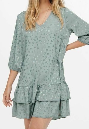 Mujer con un vestido suelto de color verde claro con un sutil estampado de leopardo, mangas de tres cuartos, escote en V y falda de volantes en capas.