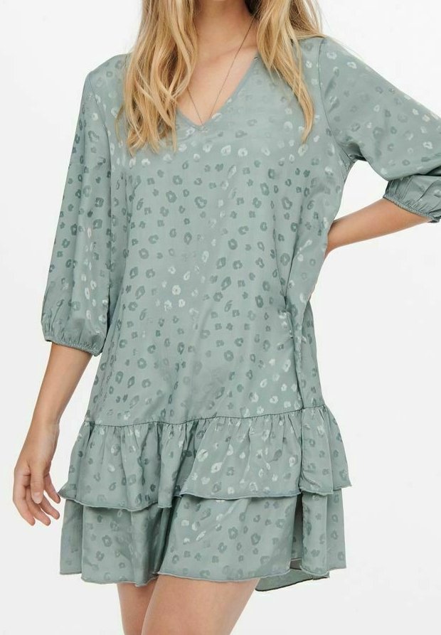 Femme portant une robe ample vert clair avec un subtil imprimé léopard, manches trois-quarts, col en V et jupe à volants superposés.
