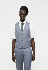 Άντρας με σκουρόχρωμο δέρμα και ντρέντλοκς φορά ανοιχτό μπλε γιλέκο και παντελόνι κοστουμιού πάνω από λευκό πουκάμισο με κοντά μανίκια και πράσινη γραβάτα, στέκεται μπροστά σε λευκό φόντο.