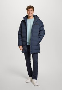 Giacca imbottita blu navy con texture trapuntata, chiusura frontale con zip e cappuccio con cordino, abbinata a una felpa azzurra chiara e pantaloni scuri.