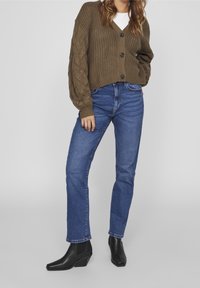 Kvinna klädd i brun kabelstickad cardigan, vit skjorta, blå raka jeans och svarta ankelboots mot en enfärgad bakgrund.