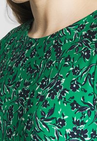 Vihreä kangas, jossa on tummansinisiä ja valkoisia kukkakuvioita. Materiaali on sileäpintainen, ja siinä on pyöreä pääntie sekä lyhyet hihat.