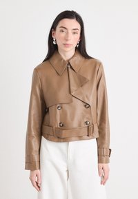 Een vrouw met lang, donker haar draagt een bruin leren dubbele-breasted jas en witte broek, poserend tegen een lichte achtergrond.