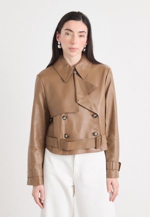 Una donna con lunghi capelli scuri indossa una giacca di pelle marrone a doppio petto e un paio di pantaloni bianchi, posando su uno sfondo chiaro.