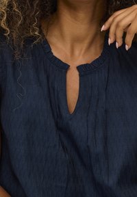 Mörkblå topp med volangutringning, nyckelhålsöppning och subtil struktur. Har plisseringar och en avslappnad passform.