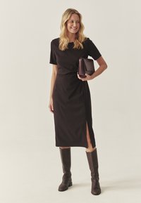 Vestido negro ajustado con mangas cortas y una abertura lateral, combinado con botas de cuero marrón hasta la rodilla y un pequeño bolso morado.