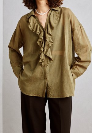 Persoon die een olijfgroene blouse met ruches en lange mouwen draagt, samen met een zwarte broek, geaccentueerd met een witte parelketting.