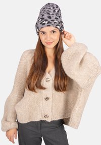 Beige gestrickte Strickjacke mit übergroßen Ärmeln und drei metallischen Knöpfen. Graue Mütze mit einem schwarz-weißen Leopardenmuster.