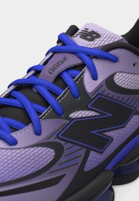 Αθλητικό παπούτσι New Balance σε μωβ και μαύρο χρώμα με μπλε κορδόνια και ετικέτα "2000" στη γλώσσα, που δείχνει υφασμάτινη υφή και λεπτομέρειες σχεδιασμού.