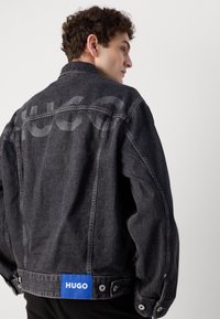 Sort denimjakke med en let falmet finish. Har et stort HUGO-logo i en lysere nuance på ryggen og et blåt mærke ved kanten.