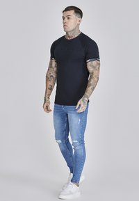 Zwarte T-shirt met korte mouwen en reliëflogo, gecombineerd met lichtblauwe versleten skinny jeans en witte sneakers. Effen achtergrond.
