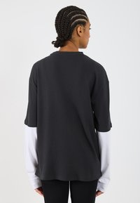 Szary, prążkowany t-shirt o luźnym kroju i opuszczonych ramionach, nałożony na długie białe rękawy, wizualna tekstura kontrastuje z gładkością.