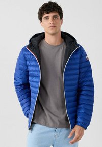 Giovane uomo che indossa una giacca a puffo blu con cappuccio, una maglietta grigia e jeans azzurri chiari, in piedi di fronte a uno sfondo chiaro e semplice.