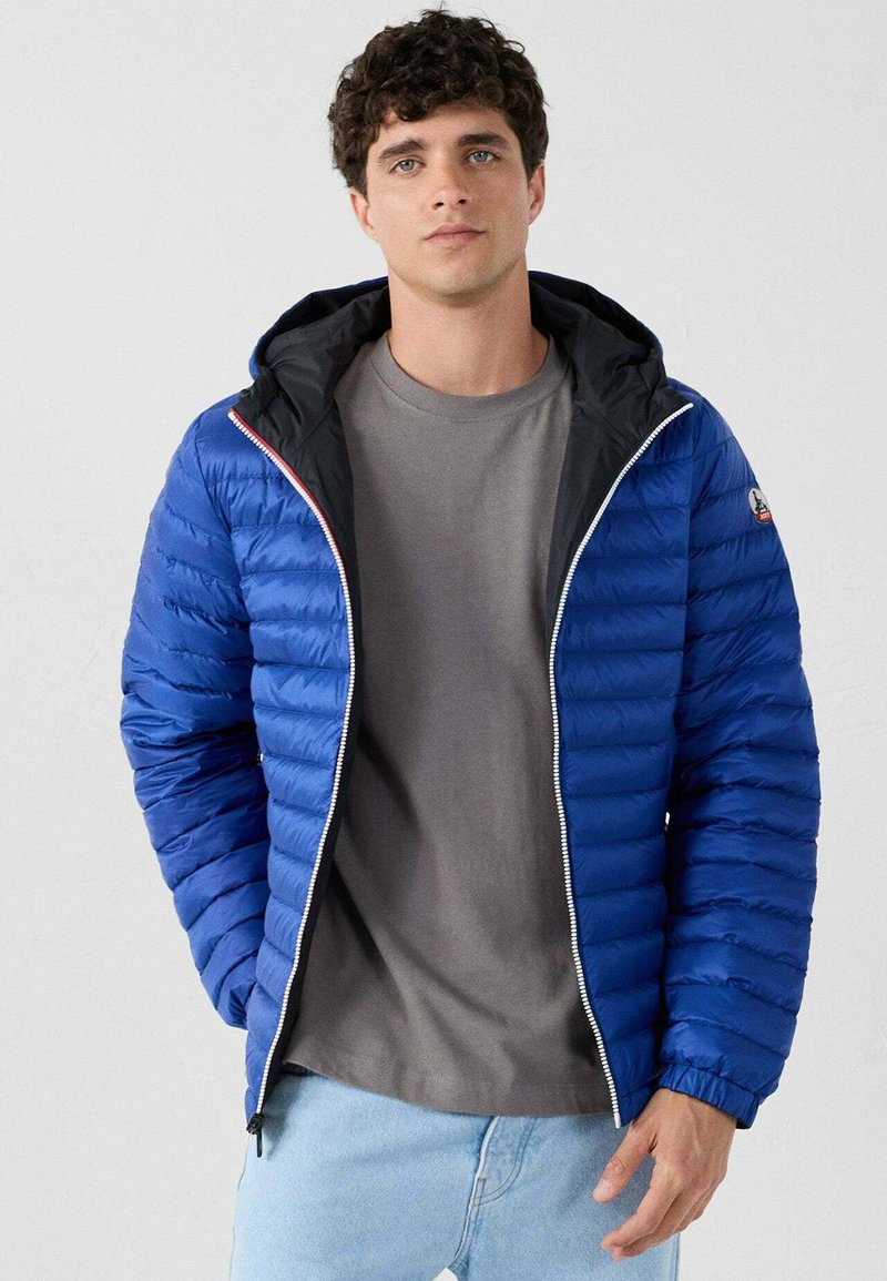 Giovane uomo che indossa una giacca a puffo blu con cappuccio, una maglietta grigia e jeans azzurri chiari, in piedi di fronte a uno sfondo chiaro e semplice.