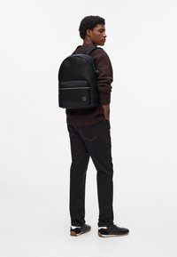 Schwarzer Leder-Rucksack mit strukturiertem Finish, vorderer Reißverschlusstasche und Logo-Akzent. Getragen mit einem braunen Pullover und schwarzen Hosen.