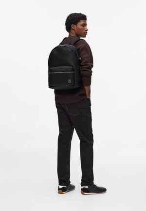 Black leather backpack with a textured finish, front zip pocket, and logo accent. Worn with a brown sweater and black pants.