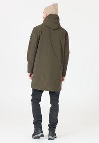 Parka impermeabile verde oliva con cappuccio, vestibilità affusolata e texture sottile. Abbinata a jeans scuri e scarpe nere robuste.