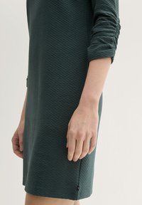 Sötétzöld, texturált ruha hosszú ujjal, bordázott mintázattal. Figyelemre méltó részlet egy apró, márkás címke a szegély közelében.