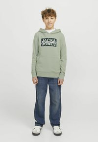 Grön huvtröja med framficka och vit grafisk text; matchas med lösa blå jeans och svarta skor med vita detaljer.