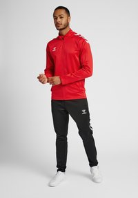 Rød zip-up sportjakke med lange ærmer, sorte bukser med sidelommer og hvide sneakers med en struktureret overflade.
