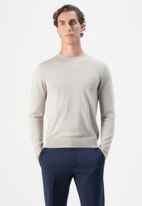 Mann mit braunem lockigem Haar, der einen hellgrauen Langarmpullover und eine marineblaue Hose trägt, steht mit den Händen in den Taschen vor einem weißen Hintergrund.