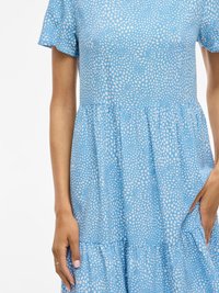 Jasnoniebieska sukienka z materiału w biały wzór w kropki. Posiada dopasowaną górę, krótkie rękawy i rozkloszowaną, wielowarstwową spódnicę.
